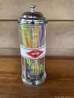 Full view of vintage Coca Cola soda straw dispenser showing clear glass cylinder, Coca Cola logo and colorful plastic straws inside.