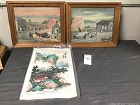 Three artworks displayed on a table, two framed folk style paintings and one Asian landscape fabric art in protective plastic sleeve.