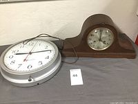 Large silver metal-framed round wall clock and a wooden mantle clock with rounded top sitting together on table