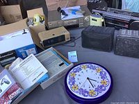 Photo showing the ceramic clock behind a digital clock radio, boxed CyberHome CD player with owner's guide, assorted cables, and remotes