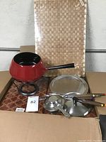 Red fondue pot with burner, two metal lids, six wooden-handled forks, placemats, and unopened beige woven tablecloths