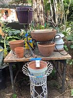 Full view of all items displayed on and around a rusty metal table including several terracotta pots, a purple ceramic pot on wrought iron stand, white decorative metal pot stand with a planter, yellow enamel pitcher.