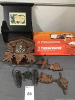 Overall view of cuckoo clock parts including clock face, broken wooden leaves, chains and weights, boxed Thermophore heating pad, and black wooden wall hanging with decorative tile and hooks.