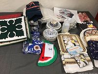 Overview of various kitchen textiles including Norman Rockwell pot holder and dish towel set, folded table coverings, potholders crocheted and printed, and multiple teacups with saucers.