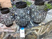 Three glass and metal lanterns with black geometric metal framework displayed on a stone ledge outdoors.