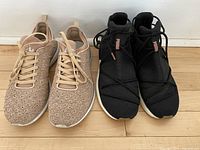 Two pairs of ladies running shoes side by side on wooden floor: beige Athletic Propulsion Labs knit shoes and black PUMA lace and strap shoes.