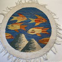 Full view of oval hand woven wool wall hanging or table mat showing fish motif on blue background with cream border and fringe.