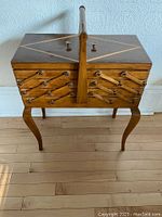 Front angled view of the wooden sewing box showing the diamond inlay on top, hinged layers with accordion-style extension arms and original brass handles, positioned on gently curved legs on a wooden floor.