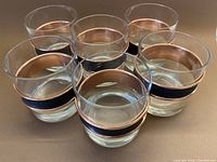 Set of six clear lowball glasses with removable copper and pleather grip bands arranged on a brown surface