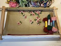 Wooden rectangular embroidery frame with partially completed pink floral needlepoint on canvas and several skeins of colored wool yarn on top and to the side of the frame.