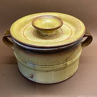 Front view of the signed clay casserole pot showing the lid, knob handle, and side handles with mustard yellow glaze and cinnamon highlights.
