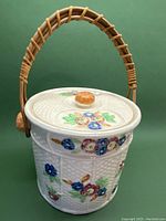 Photo of vintage hand-painted ceramic cookie jar with textured surface, floral designs, and bent bamboo handle on green background.
