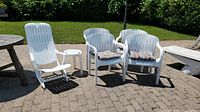 Four matching white plastic outdoor chairs with beige cushions and one taller white plastic lounge chair without cushion on a patio.