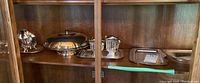 Multiple plated copper serveware items: sugar bowl, cake dish with cover, creamer pitcher on tray, large rectangular tray on wooden shelf