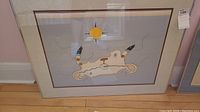 Framed print of First Nations artwork with animal figure and yellow sun, shown resting on a wood floor from a side angle.