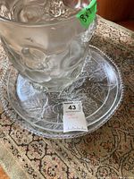 Glass vase with embossed cherub or angel faces and wings, standing on two clear glass plates with decorative floral/leaf patterns, on a patterned carpet.