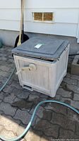 Plastic hose reel featuring a hinged black lid and crank handle on the front. Beige in color placed outdoors on stone pavement.