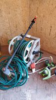 Overall view of garden hose on reel, brush attachment, and sprinklers on wooden floor against particle board wall.