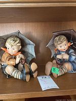 Two Hummel figurines of a boy and a girl sitting with umbrellas, side view.