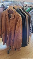 Photo showing six jackets hanging on rack; tan leather jacket in front with visible pockets and button front
