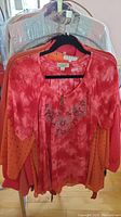 First photo shows the red tie-dye blouse with floral embroidery and small buttons on the chest, hanging on a rack with other tops behind it.