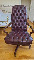 Full front view of brown leather tufted office chair with wooden armrests and wheeled base showing riveted and puckered upholstery finish.