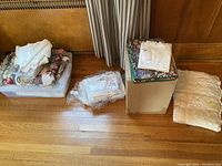 Overview photo of the lot showing folded linens in containers and boxes placed on the floor, including crocheted and floral patterned linens.