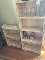 Two wooden shelf units side by side, one taller with 4 tiers and one shorter with 2 tiers, both show decorative carved edges and some contents on the shelves (glasses and dolls)