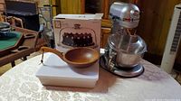 KitchenAid professional mixer and accompanying kitchen items on table: wooden bowl with handle, white rectangular ceramic baking dish, boxed glass cake plate.