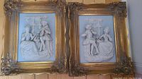 Two framed dimensional art plaques showing detailed relief scenes of a man and woman in period costume in a garden setting with decorative golden frames.