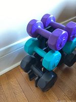 Photo showing colorful hand weights stacked on the black plastic stand against a wall and hardwood floor.