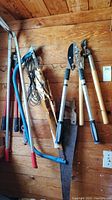 Photo showing multiple yard tools hanging on wooden wall including loppers, bolt cutter, saw, pruners with handles of wood, metal and rubber grips.