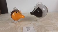 Photo of two glass fish figurines, one orange and one with black, white, and brown color sections, placed on a cloth surface.