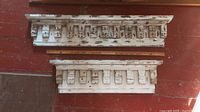 Wide view of two carved wooden architectural elements with distressed white paint, placed on red wooden floor, showing size comparison with measuring sticks.