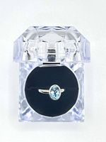 Top-down view of blue Swiss topaz ring inside clear acrylic ring box showcasing the oval blue gemstone and silver band.