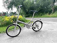 Full side view of the silver Next Compact folding bike on pavement with foliage background