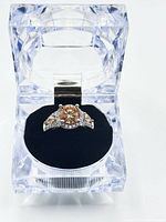 Front angle of the padparadscha sapphire ring in a clear acrylic box showing the size and color of the main stone and side topaz.