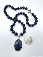 Full view of lapis lazuli necklace with oval pendant and a coin for scale to show bead size and pendant dimensions.