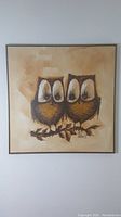 Photo of framed square oil painting showing two stylized owls on a branch with warm beige background, signed "Travis"