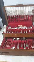 Full open wooden case with red velvet lining holding the complete set of cutlery pieces distributed across three layers and compartments.