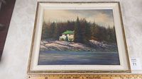 Framed oil painting showing lakeside cottage scene with surrounding pine trees and rocky shore.