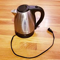 Side view of Hamilton Beach electric tea kettle on wooden floor, showing water level indicator, handle, lid, and power cord.