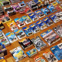 First photo shows a collection of approximately 60+ 1:64 scale diecast cars on original blister cards laid out on wooden floor. Mostly Hot Wheels blue cards with some Matchbox and boxed sets.