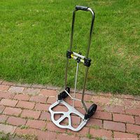 Fold-up two-wheel push cart standing with base extended on brick surface with grass background.