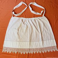 Full view of natural/off-white cotton apron with shoulder straps and gathered waist, laid flat on orange background.