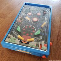 Blue vintage Tomy toy pinball machine with clear plastic playfield cover, red ball inside, red and yellow flippers and control buttons on bottom right