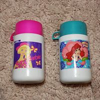 Front view of two vintage plastic thermoses with colorful Barbie and The Little Mermaid illustrations, placed on carpet.