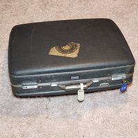 Closed view of vintage black hard plastic suitcase with partial label residue and silver hardware resting on carpet.
