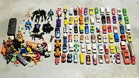 Overview photo showing over 70 diecast cars arranged in rows with action figures and toys to the left side. Various colors and vehicle types visible.