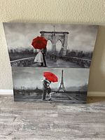 Two canvas wall art pieces stacked against a wall showing romantic couples with red umbrellas on city backgrounds including Brooklyn Bridge and Eiffel Tower.